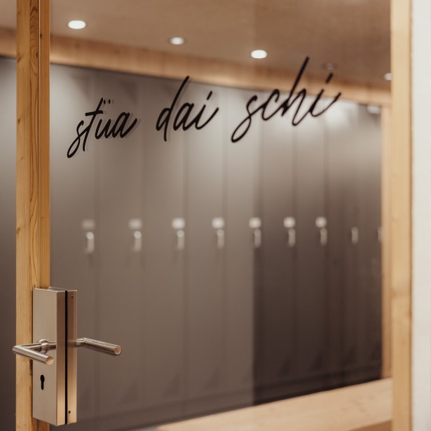 Porta della sala d'asciugatura con la scritta ladina stüa dai schi. Sullo sfondo sono visibili i contorni sfocati dei singoli armadietti.