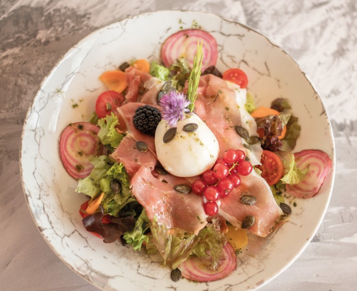 Piatto ben presentato visto dall'alto, con insalata, burrata, ribes, cipolle, prosciutto e more.