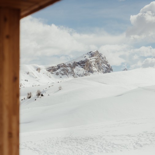 View through a window with a wood frame onto the vast, solitary snow-covered mountain landscape. Dolomite peaks under a cloudy sky.