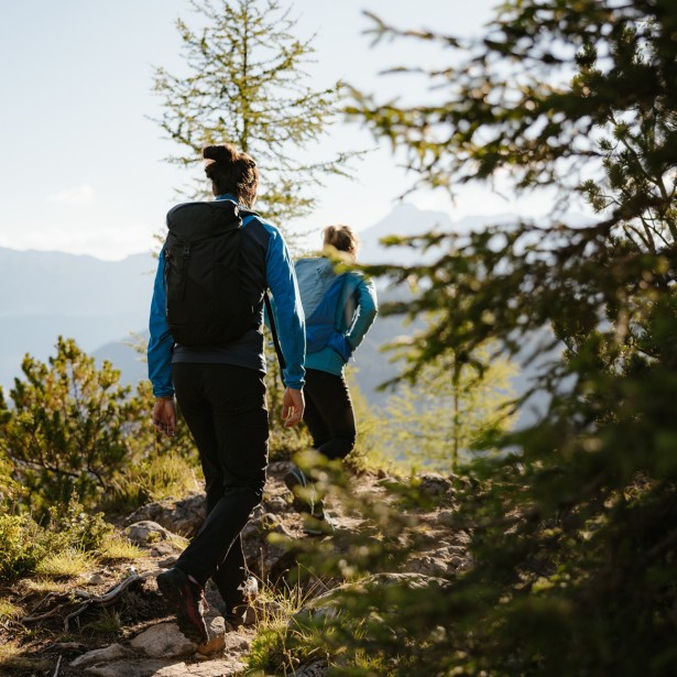 Due escursionisti su un sentiero di montagna sassoso, circondati da una foresta verde, con vista su un vasto panorama montano.