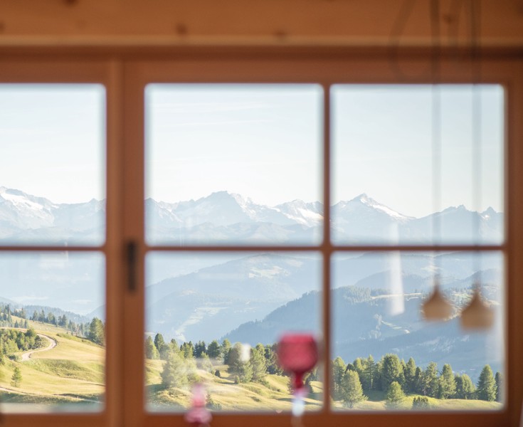 La vista attraverso una tradizionale finestra alpina con telaio in legno rivela che solo natura incontaminata circonda il rifugio alpino Pralongià: colline erbose, ampie conifere e alte montagne.