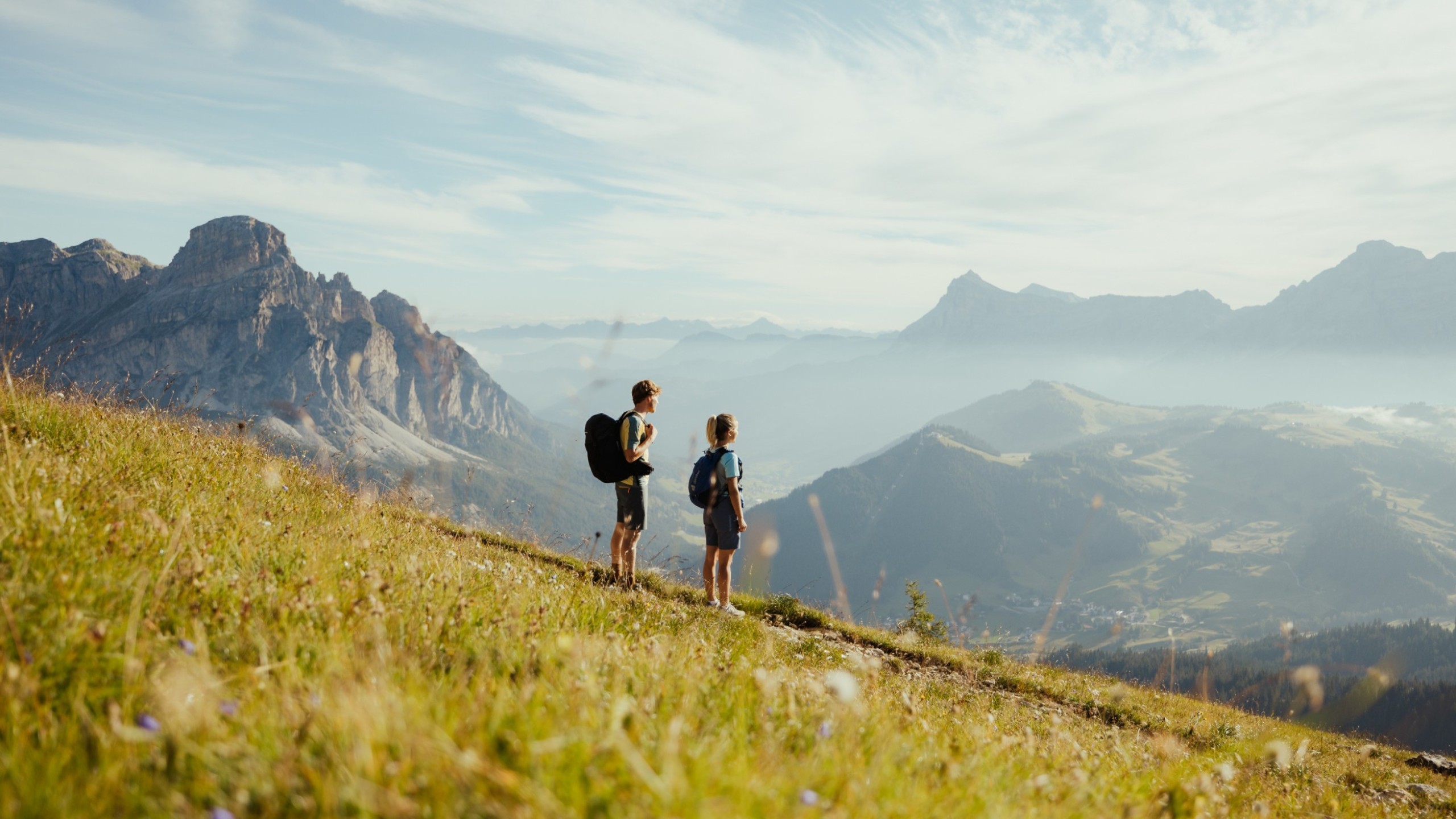 Due escursionisti sono su un sentiero, circondati da un prato fiorito e guardano il panorama montano mozzafiato in lontananza.