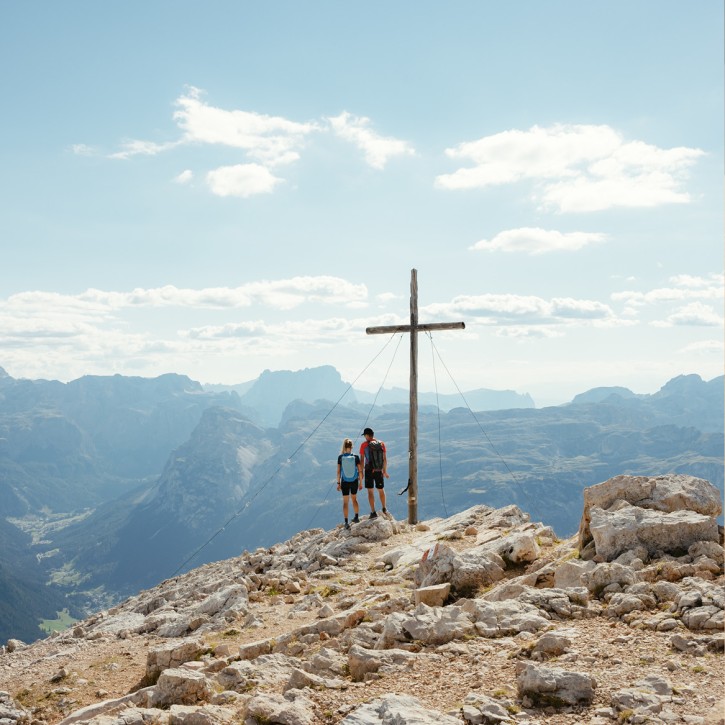 Una donna e un uomo sono in piedi presso la croce di vetta e guardano il vasto panorama montano in lontananza sotto un cielo sereno.