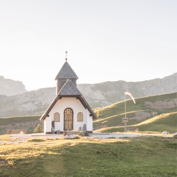 Small, sun-drenched chapel surrounded by a sparse meadow and alpine landscape.