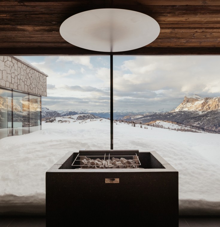 Moderner Saunaofen mit Steinen vor einer großen Fensterfront, die einen Panoramablick auf die verschneite Berglandschaft der Dolomiten bietet.