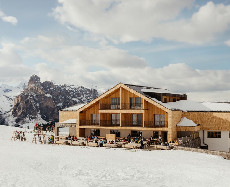 Il Rifugio Alpino Pralongià con la sua facciata in legno e la terrazza vista fronte in inverno, sullo sfondo la nota cima dolomitica del Sassongher.