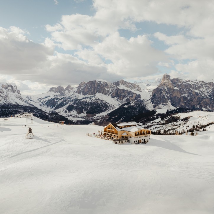 Il Rifugio Alpino Pralongià a volo d'uccello. Sorge solitario su un ampio prato alpino innevato. Sullo sfondo, le Dolomiti sotto il cielo nuvoloso.