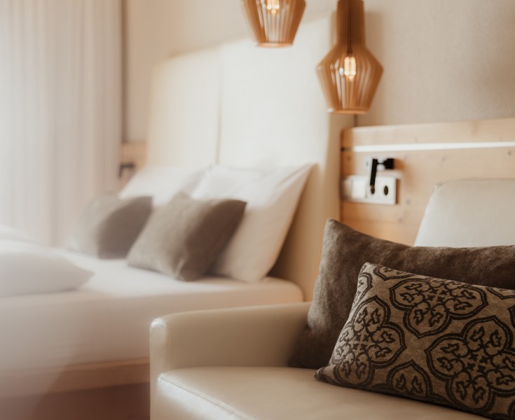 Due cuscini morbidi e vellutati con un motivo sono appoggiati su una poltrona di pelle, e dietro si trova un accogliente letto matrimoniale con una testiera alta.