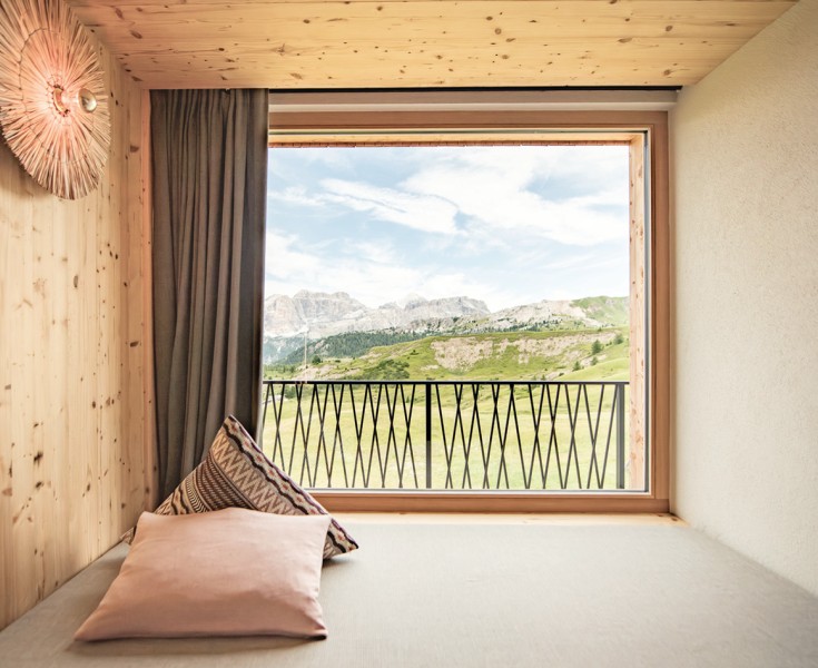 Camera accogliente e luminosa con pareti in legno, un letto matrimoniale e una grande vetrata che lascia spazio alla vista sul paesaggio montano.