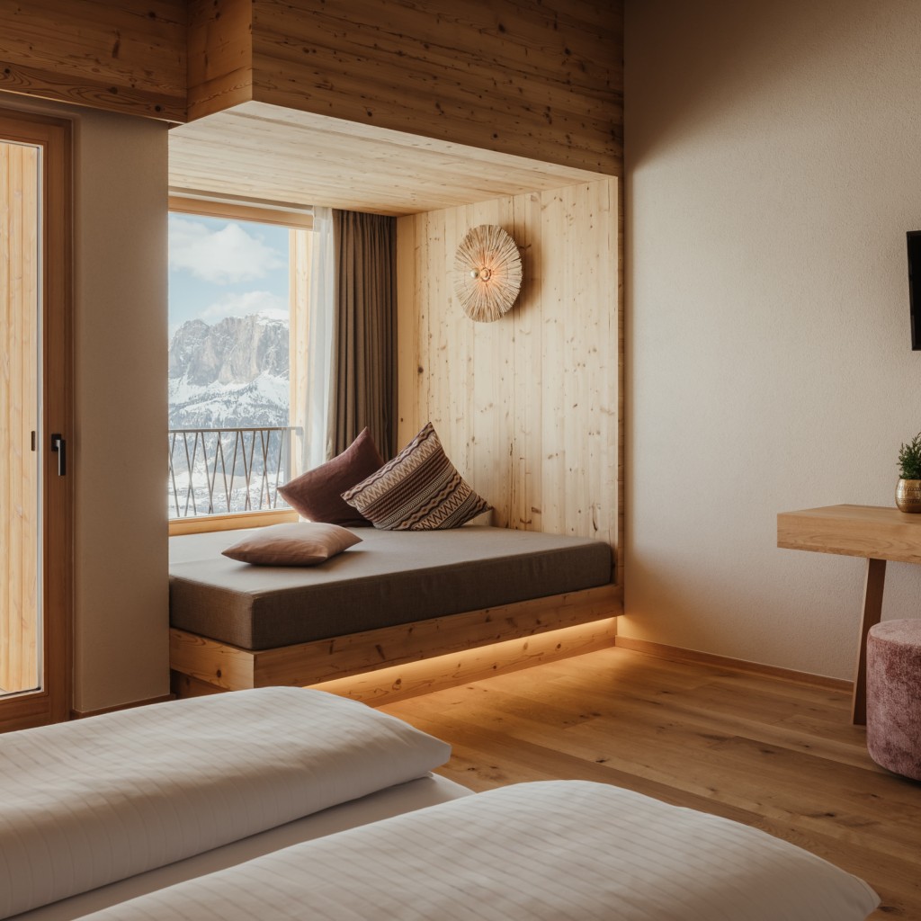 Camera da letto luminosa e tranquilla con pavimento in legno vero, rivestimento in legno massello e un comodo spazio relax direttamente alla finestra con vista sulle Dolomiti.