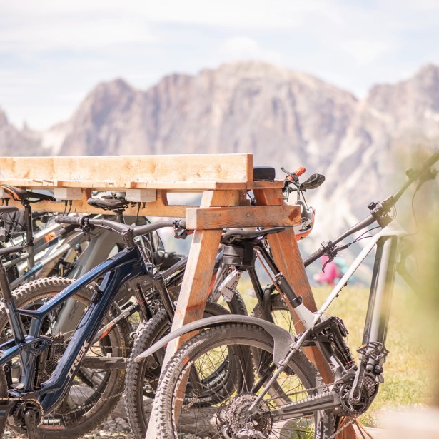 Diverse mountain bike sono appoggiate a una rastrelliera di legno all'aperto. Sullo sfondo si trovano alcune montagne delle Dolomiti.