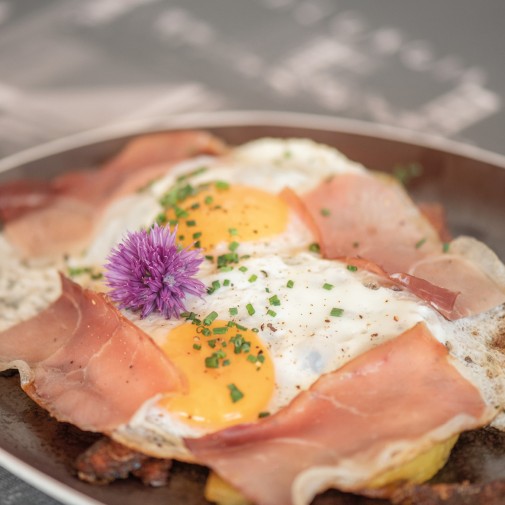 Una padella con uovo all'occhio di bue e fette di speck, presentata con cura. Il piatto è guarnito con erba cipollina fresca e un fiore decorativo commestibile.