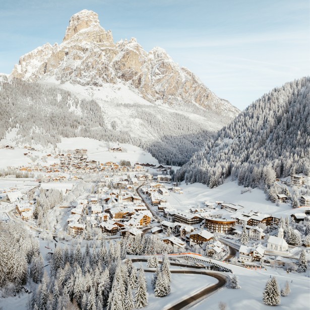The snowy village of Corvara at the foot of the snow-covered, prominent Dolomite mountain Sassongher, located in the middle of the Dolomites and surrounded by forest.