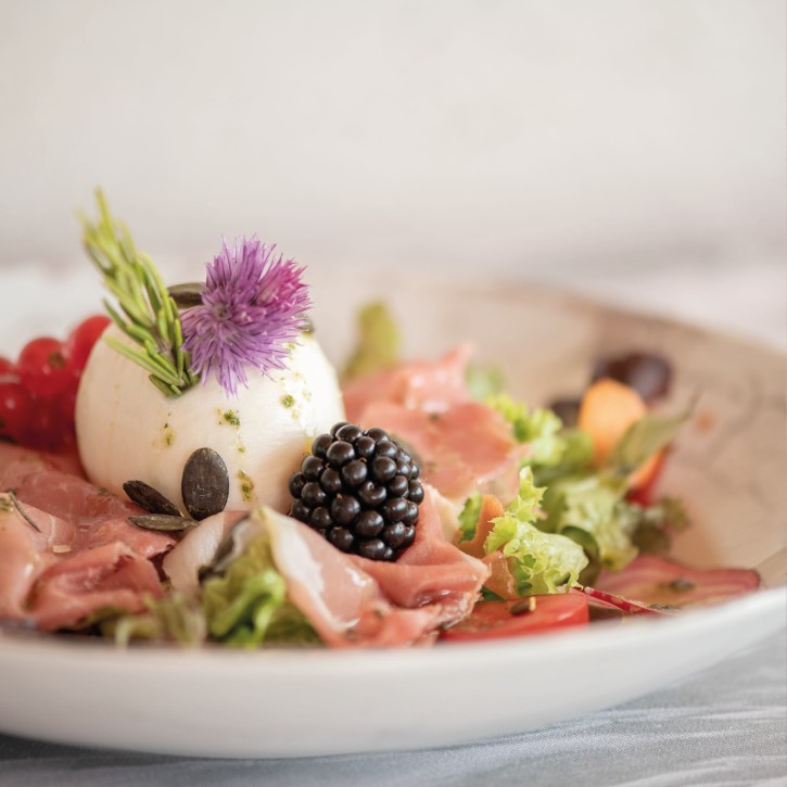 Ansprechend angerichteter Teller mit grünem Blattsalat, Burrata, Brombeeren, roten Johannisbeeren, einer essbaren Blüte und Schinken.