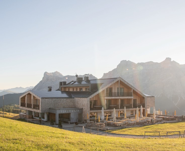 Il Rifugio Alpino Pralongià in estate, al centro di un alpeggio verde, con la terrazza vista fronte e le Dolomiti sullo sfondo.