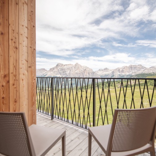 Balkon mit zwei Stühlen als Sitzgelegenheit in der frischen Bergluft und einem Metallgeländer mit Rautenmuster. Der Blick fällt auf die grüne Wiese und die Berglandschaft unter bewölktem Himmel.