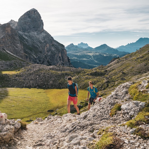 Due persone stanno camminando insieme su un piccolo sentiero sassoso in salita. Si trovano già in alto sulla montagna, da qualche parte nelle Dolomiti, e sono circondati da un impressionante paesaggio montano.