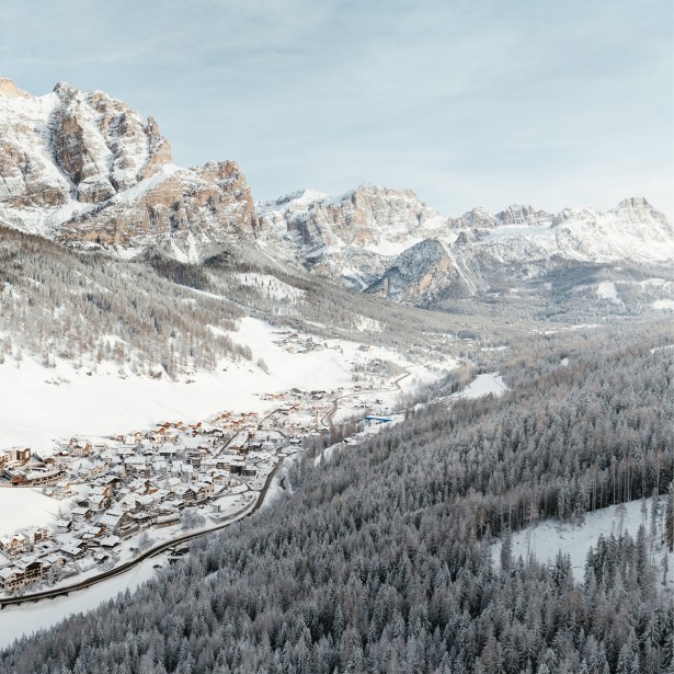 The picturesque, small snow-covered village called San Cassiano in a pristine valley viewed from a bird's-eye perspective, surrounded by equally snow-covered forests and Dolomite rocks.