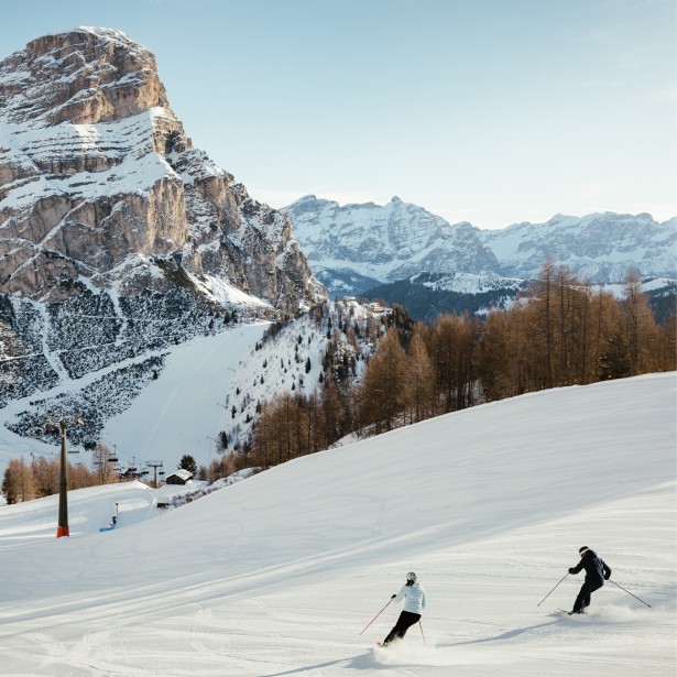 Two skiers descending a groomed slope, with breathtaking Dolomite rocks and a magnificent mountain panorama in the background.