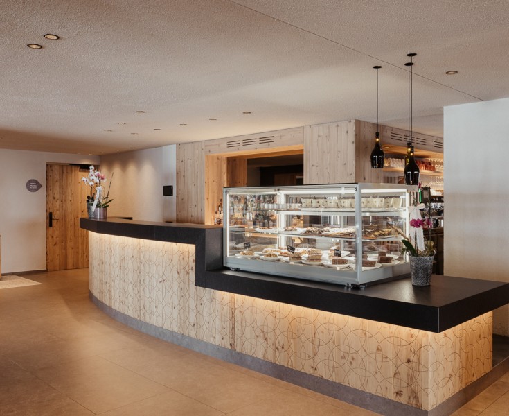 Il bancone del bar, con rivestimento in legno e motivo inciso, sul quale sono esposti vari dolci in una vetrina.