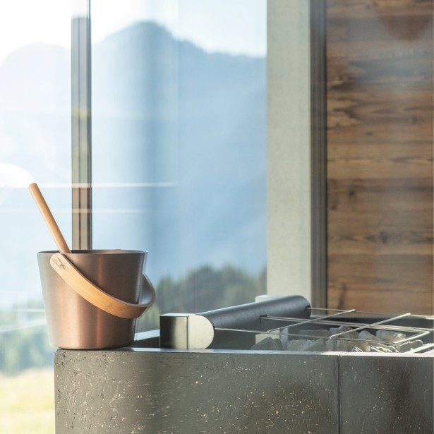 Area sauna con rivestimento in legno e una vista mozzafiato sulle Dolomiti in lontananza. Il secchio per il maestro di sauna e la prossima gettata di vapore è pronto.