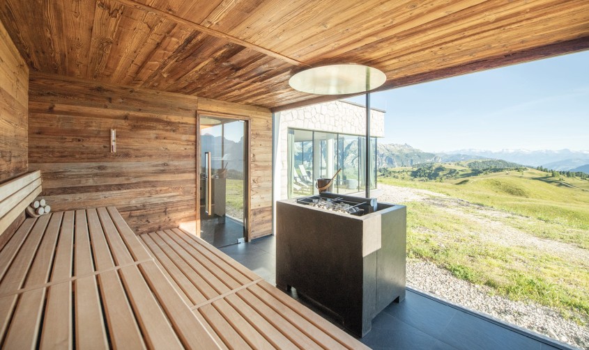 Sitzgelegenheiten aus Holz in der hellen Sauna mit viereckigem Saunaofen in der Mitte und großer Fensterfront mit Blick auf die sonnige Berglandschaft.