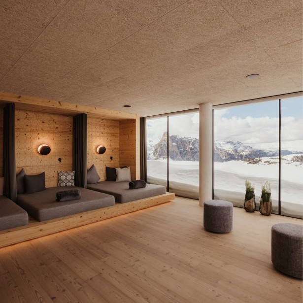 Großer, ruhiger Raum mit Sitzkissen und mehreren weichen Kissen als Ruhemöglichkeit an einer Holzwand und mit Blick durch die große Fensterfront auf die verschneite Berglandschaft.