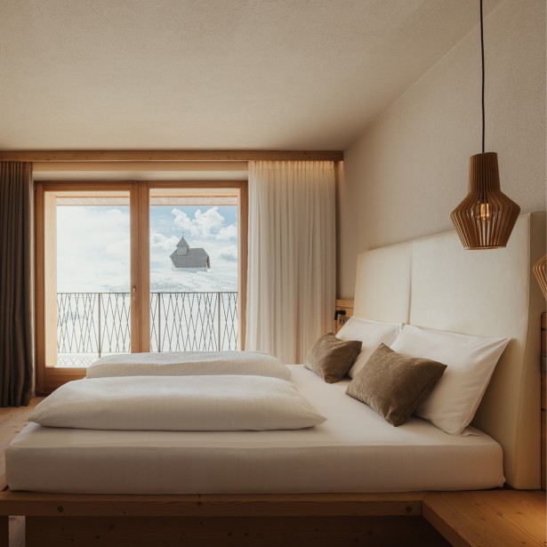 Modernly furnished bedroom with a double bed, cuddly pillows, wooden floor, calm atmosphere, and a view of a snowy mountain landscape with a chapel.