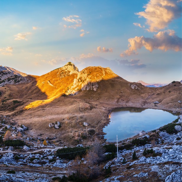 Kleiner Bergsee, umgeben von kargen Almwiesen und vielen Dolomit-Steinen. Die Sonne beleuchtet einen einzigen Gipfel, der Rest liegt im Schatten.