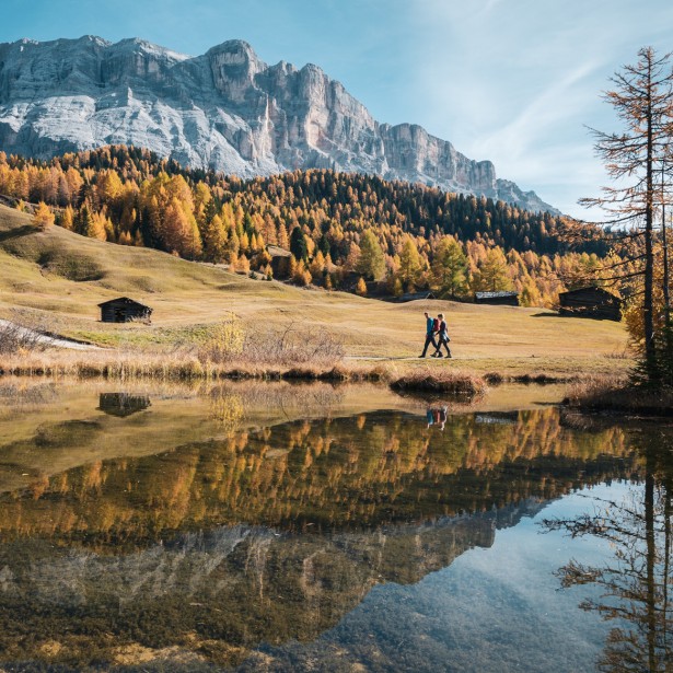 Due escursionisti camminano in autunno lungo un lago la cui superficie calma riflette le montagne circostanti e il bosco autunnale colorato.