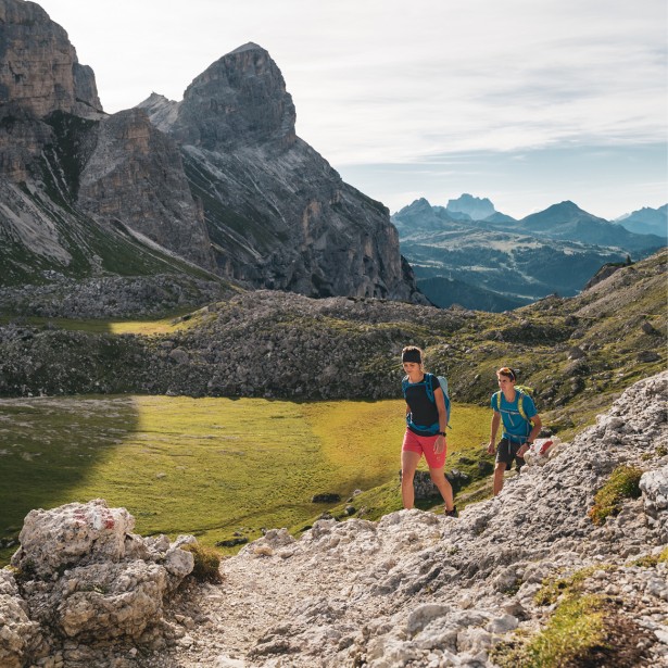 Zwei Menschen wandern gemeinsam auf einem kleinen, steinigen Weg nach oben. Sie befinden sich bereits hoch oben am Berg, irgendwo in den Dolomiten, und sind umgeben von einer beeindruckenden Berglandschaft.