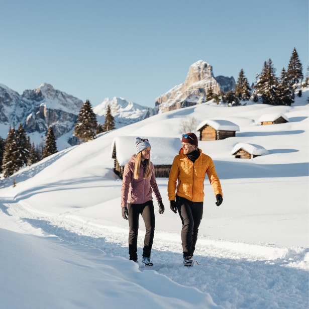 Two people in winter jackets and hats are hiking on a winter hiking trail, with snowy huts, trees, alpine meadows, and mountains in the background.