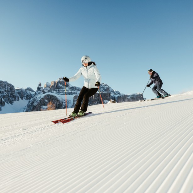 Two skiers, a woman in a white jacket with a helmet in the front, and a man in completely black attire in the back, are skiing on a freshly groomed slope in the Dolomites.