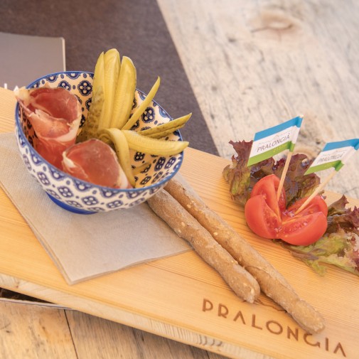 Una ciotola fonda con speck e fette di cetriolo è su un tagliere di legno. Accanto si trovano insalata, pomodori e due grissini spessi. Due bandierine con la scritta “Pralongiá” sono infilate in un pomodorino ciliegino.