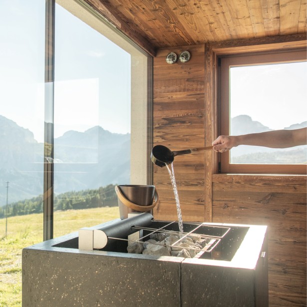 Ein Saunameister gießt mit einem Schöpfer Wasser auf die heißen Steine im Saunaofen. Große Fensterfront und Blick auf die Dolomiten.