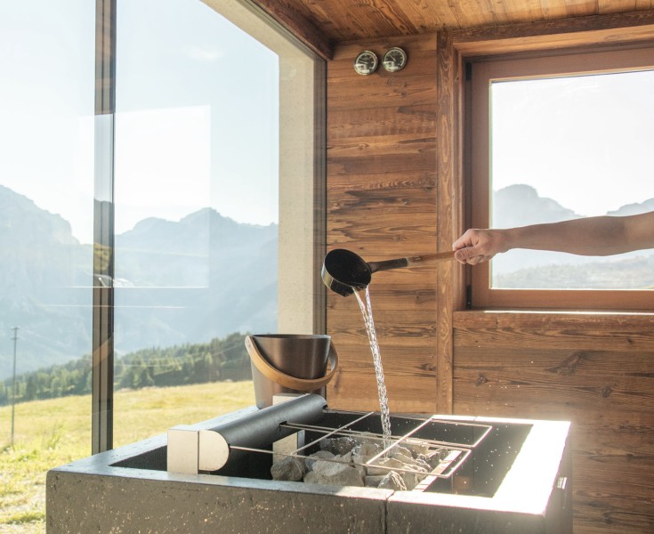 Un maestro di sauna versa acqua sulle pietre calde nella stufa della sauna con un mestolo. Ampia vetrata e vista sulle Dolomiti.