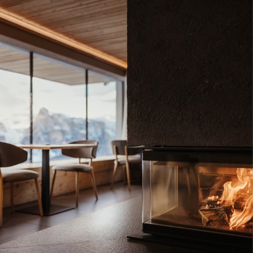 In einem schwarzen Ofen brennt ein Feuer und sorgt für eine gemütliche Atmosphäre. Dahinter Esstische neben einer langen und hohen Fensterfront, die den Blick auf die atemberaubende Berglandschaft freigibt.