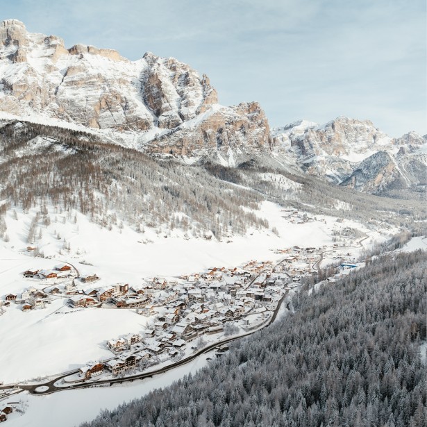 A picturesque, small snow-covered village in a pristine valley viewed from a bird's-eye perspective, surrounded by equally snow-covered forests and Dolomite rocks.