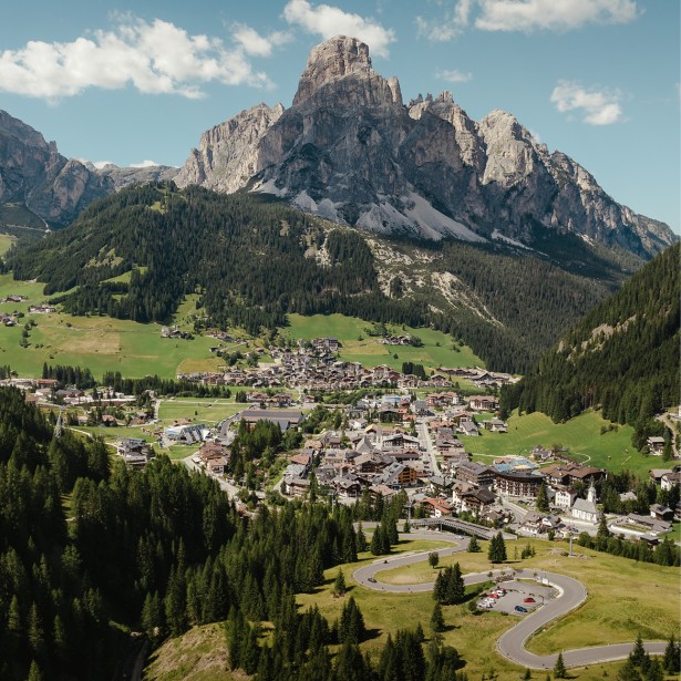  Das Dorf Corvara im Sommer aus der Vogelperspektive, umgeben von grünen Wiesen und dichten Wäldern, im Hintergrund der markante Fels des Berges namens Sassongher.