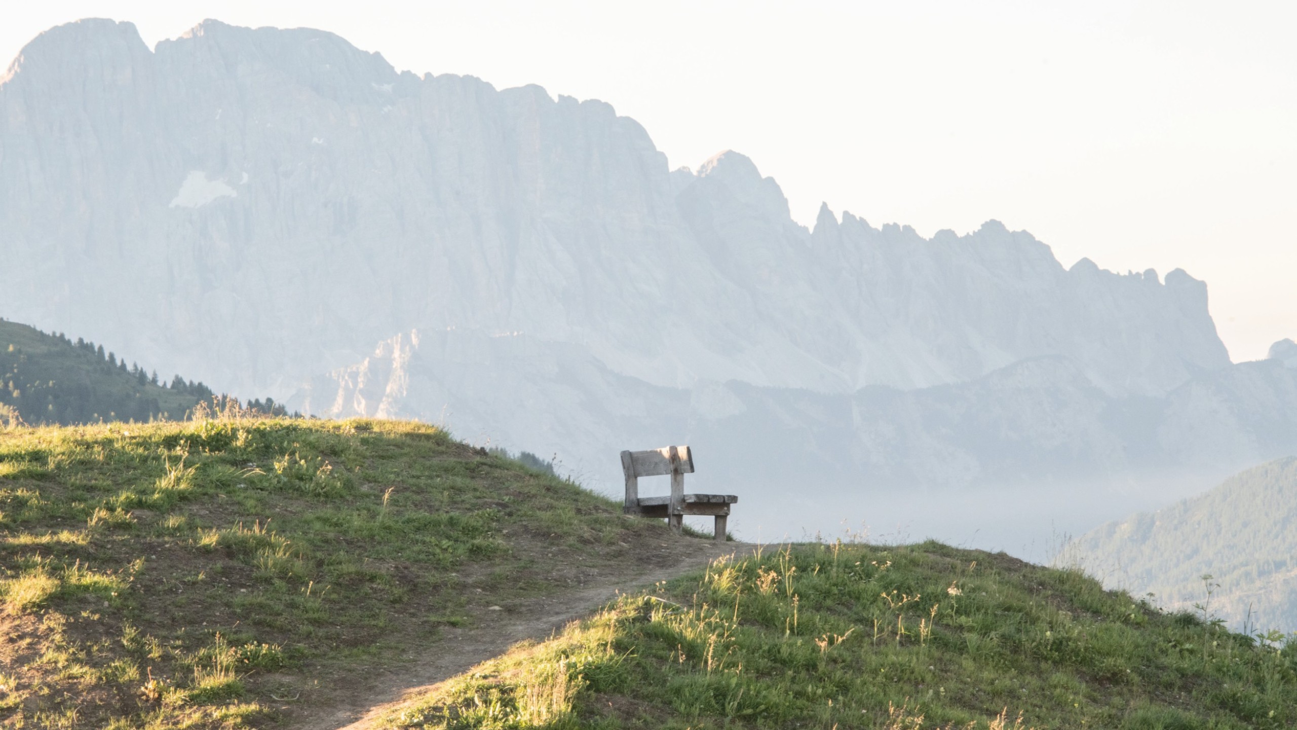 Panchina di legno solitaria su un colle erboso al sole mattutino, con una splendida vista sulle Dolomiti avvolte nella nebbia sullo sfondo.