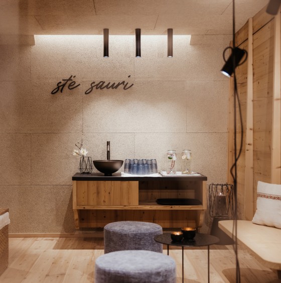 Gemütlicher Ruheraum im Wellnessbereich mit holzverkleideten Wänden. Wandregal mit rundem Waschbecken und Gläser für die Erfrischung zwischen den Saunagängen. Zwei gemütliche Hocker, ein kleiner schwarzer Tisch und der ladinische Schriftzug sté sauri an der Wand.