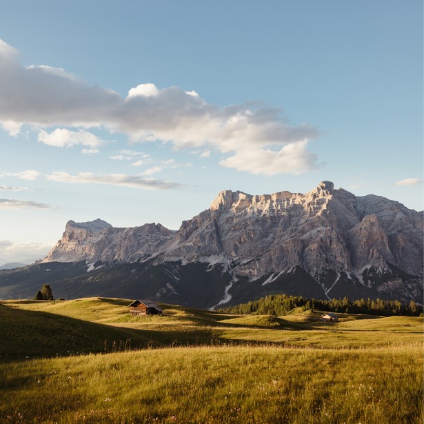 Weite grüne Bergwiesen mit einzelnen Holzhütten vor massiven Bergen unter blauem Himmel mit Wolken.