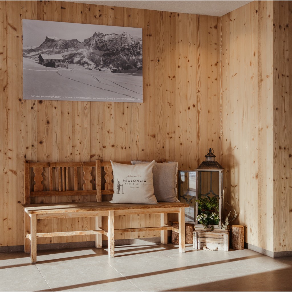 Comoda panca tradizionale in legno con due cuscini davanti a una parete con rivestimento in legno, sopra la quale è appesa una foto panoramica in bianco e nero del vecchio rifugio alpino Pralongià.