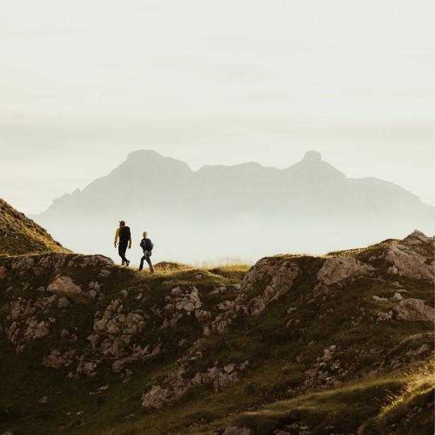 Zwei Menschen mit Rucksäcken wandern auf einem felsigem Bergrücken, im Hintergrund die Silhouette eines Berges in den Dolomiten.