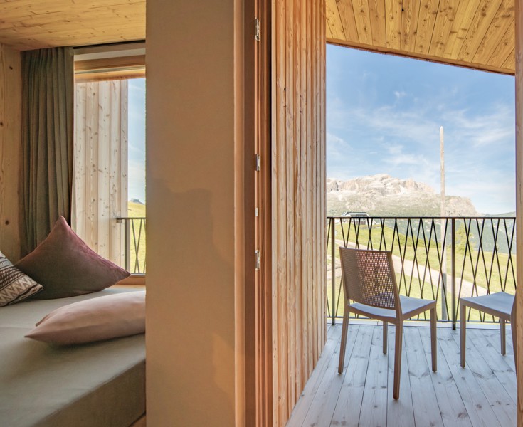 Una camera da letto accogliente e semplice con un letto e cuscini a sinistra. Un muro separa la zona notte dal balcone a destra, dove ci sono due sedie e si gode la vista sul paesaggio soleggiato.