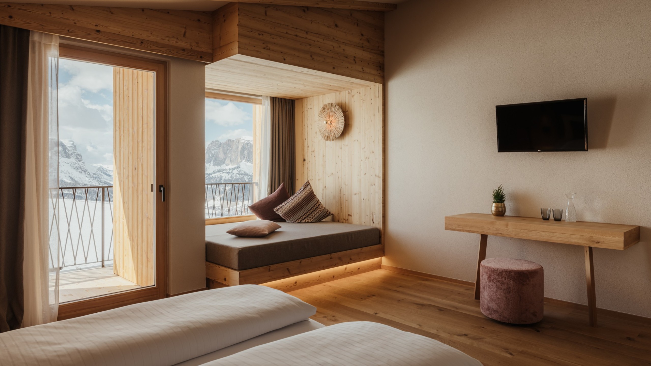 Camera da letto luminosa e tranquilla con pavimento in legno vero, rivestimento in legno massello e un comodo spazio relax direttamente alla finestra con vista sulle Dolomiti.