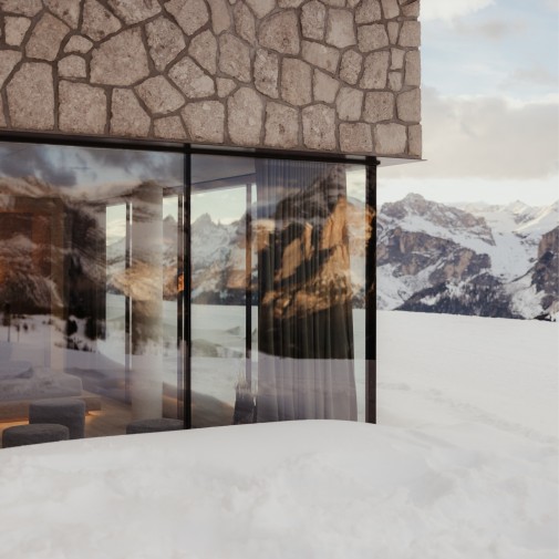 Eine der vielen großen Fensterfronten des Berggasthauses Pralongià, umgeben von Schnee. Der Blick fällt auf schneebedeckte Berge und den Innenraum mit Vorhängen und Sitzmöbeln.