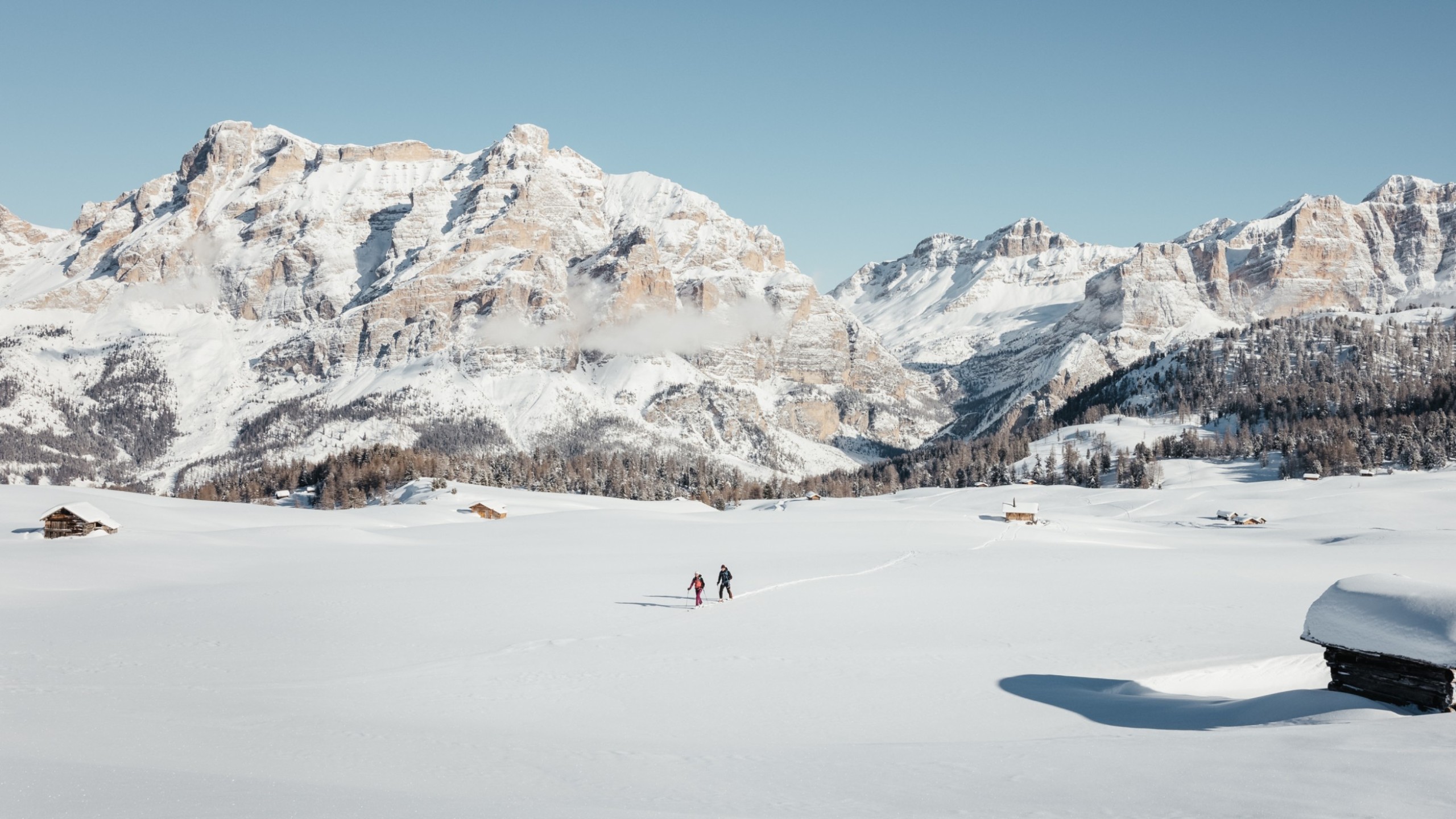 Two people are wading through a solitary and vast, sun-drenched and snow-covered alpine meadow, on which two small wooden huts are located. In the background, the snow-covered Dolomites and a coniferous forest at their base.