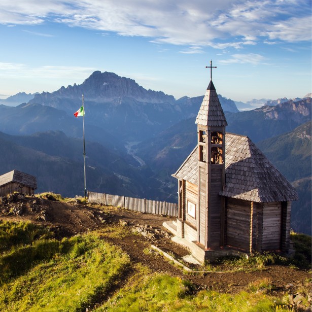 Una piccola cappella antica con facciata in legno si trova su un poggio erboso rialzato. Sullo sfondo si vede il vasto panorama montano.
