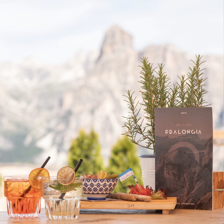 Due bevande con fette di limone e arancia e cannucce, insieme a un tagliere di legno con piccoli stuzzichini e accanto il menu sul tavolo. Sullo sfondo si intravedono le montagne, sfocate.